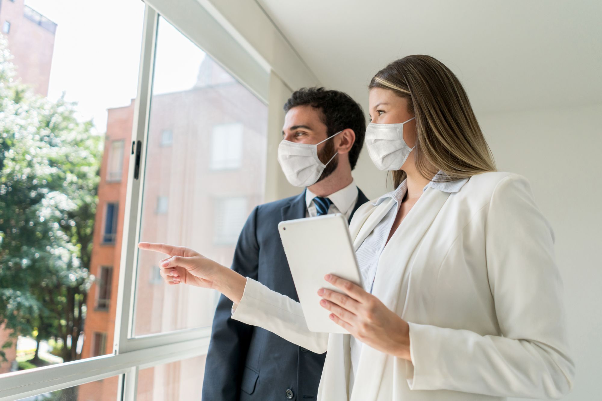 Beautiful real estate agent pointing at the view to customer both looking very cheerful but wearing protective face masks - Coronavirus concepts